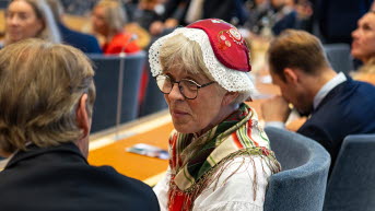 En kvinna i folkdräkt och mössa vänder sig mot en person som syns bakifrån. De båda sitter i riksdagens kammare.