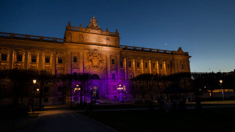Kvällsljus på Riksdagshusets exteriör. Mitten av byggnaden är upplyst med en lila färg.