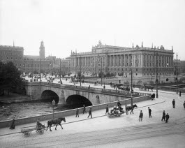 Svartvit bild på riksdagshuset 1905. Människor som går och som kör häst med vagn syns i bild.