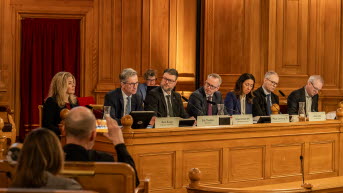 Sju personer sitter vid ett podium med träpanel bakom. I förgrunden syns några åhörare, varav en räcker upp handen. 