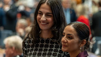 Två kvinnor ler och tittar till vänster i bilden. Bakom dem syns flera andra människor i oskärpa.