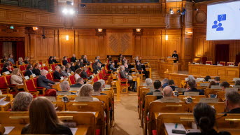 Stor publik i sal som lyssnar på en person i talarstolen längst bort. En powerpoint-presentation syns ovanför talaren.