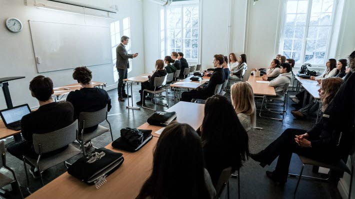 Ett klassrum fullt med elever i gymnasieåldern. Eleverna syns bakifrån och längst bort står en man i kostym.