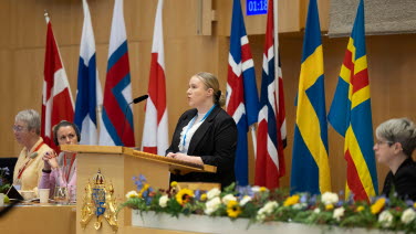 Kvinna står i talarstolen med nordiska flaggor bakom sig.