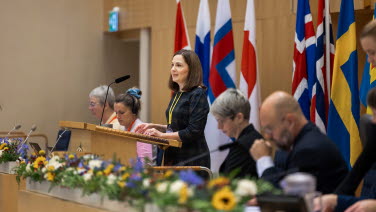 Kvinna står i talarstolen vid talmanspodiet. Vid sidan av henne sitter tjänstemän.