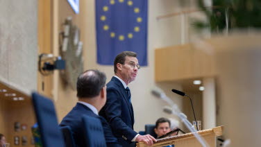 Statsministern står i talarstolen vid talmanspodiet med EU-flaggan i bakgrunden.,