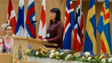 Kvinna står i talarstolen i kammaren med flaggor i bakgrunden.