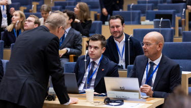 En delegat står böjd över två andra delegater som sitter i sina bänkar i kammaren.