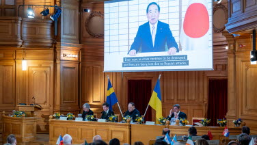 A man speaks via a big screen in the former Second Chamber. The image shows the Japanese flag.