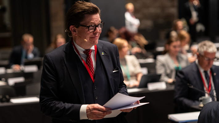 Talman Andreas Norlén går genom en sal där människor syns sitta vid bänkar i bakgrunden. Han har några papper i händerna.