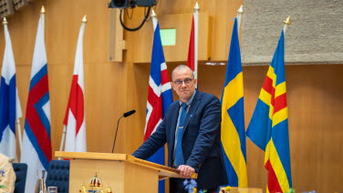 Man står i talarstolen med flaggor i bakgrunden.