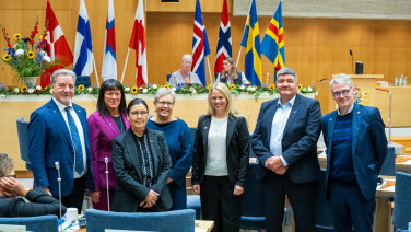 Sju personer står framför talmanspodiet i kammaren. I bakgrunden de nordiska flaggorna.