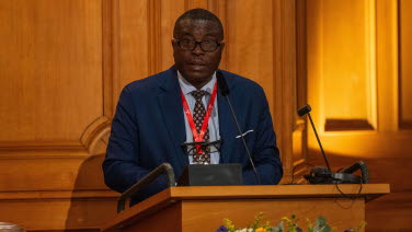 A man delivers a speech in the former Second Chamber. He wears a red lanyard around his neck.