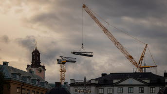 Två lyftkranar i motljus mot en mörk molnig himmel. Storkyrkans torn syns i bild.