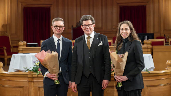 Vinnarna av uppsatstävlingen 2025 tillsammans med talmannen med varsin bukett framför ett podium i en sal med träpaneler.