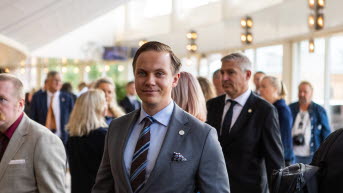 En man i grå kostym och slips står bland många andra personer i riksdagens kammarfoajé. Han ler mot kameran.