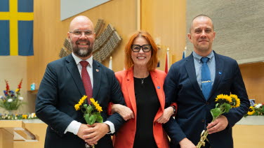 Tre personer står framme vid podiet i Andrakammarsalen.