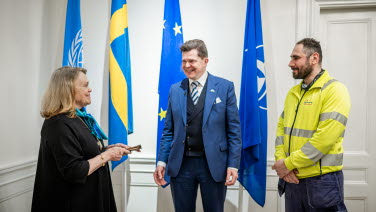 En kvinna och två män pratar leende med varandra. Kvinnan håller en plånbok i handen.