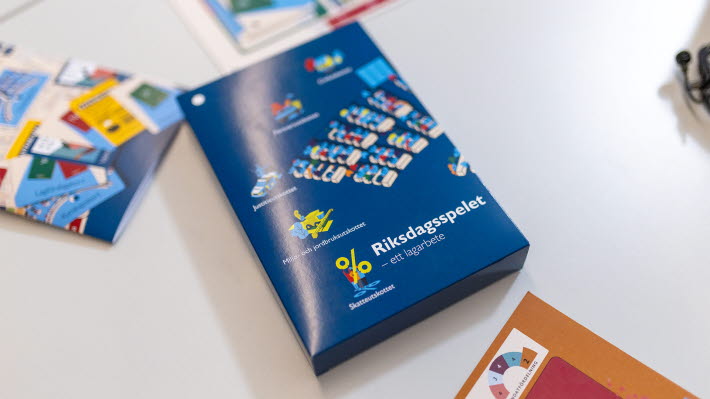 En färgglad ask som det står Riksdagsspelet på. Vid sidan skymtar ett lika färglatt häfte och en spelplan.