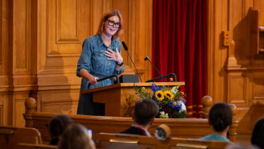 En kvinna står i talarstolen. Blommor i förgrunden. 