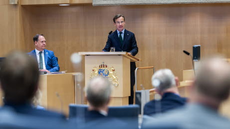 Statsministern står i talarstolen. Till vänster förste vice talman. I förgrunden sitter riksdagsledamöter och lyssnar.
