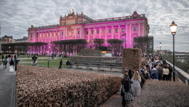 Fasaden på Riksdagshuset östra är upplyst i lila färg. Folk står i kö för att komma in.