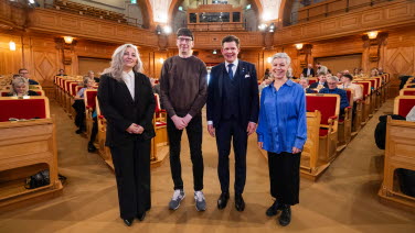 Fyra personer står och tittar in i kameran i riksdagens förstakammarsal.