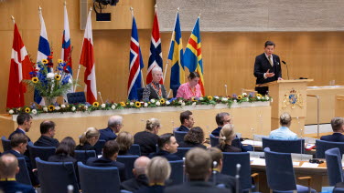 Talmannen står i talarstolen med flaggor i bakgrunden. I förgrunden ser man ledamöter som sitter och lyssnar.