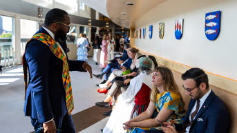 Flera personer sitter på en lång soffa. I förgrunden talar några personer med varandra. De flesta har festliga färglada kläder. På väggen bakom soffan hänger en lång rad  vapensköldar.