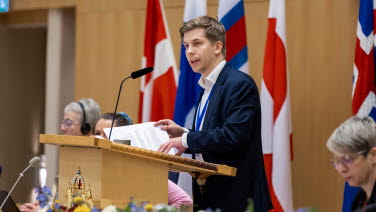 Man står i talarstolen med flaggor i bakgrunden.