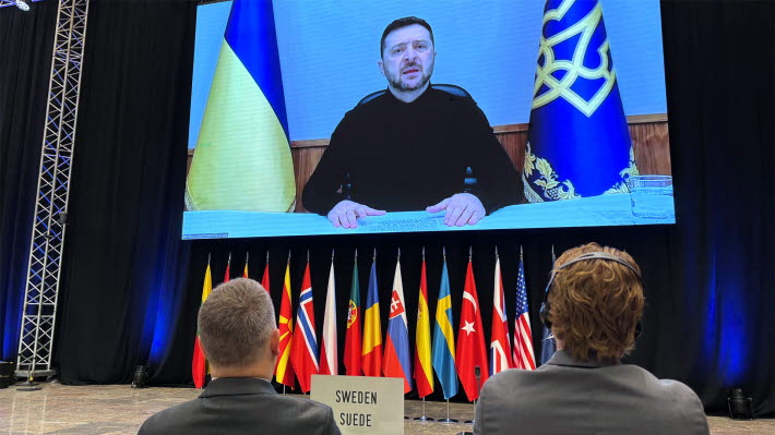 Två män i kavaj syns bakifrån. De tittar på en stor skärm där Volodymyr Zelenskyj syns. Framför männen finns en skylt: "Suede, Sweden"