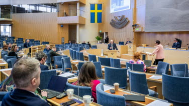 Två personer står i var sin talarstol i kammaren. Ledamöter lyssnar i sina bänkar.