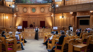 En skådespelare med kläder från början av 1900-talet berättar om riksdagen och riksdagens arbete i Andrakammarsalen.