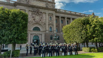 En uniformsklädd orkester står framför Östra riksdagshuset mellan två gröna träd. 