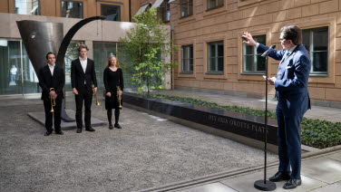 Talmannen talar i mikrofon och vänder sig till tre musiker som står framför en skulptur.