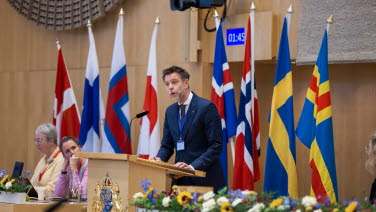 Man från norge står i talarstolen med nordiska flaggor i bakgrunden.